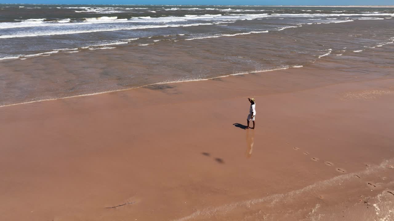 Man in white clothes walks on a curved sandbar beside muddy Parnaíba River waters in Araioses, Brazil. Aerial follow shot
