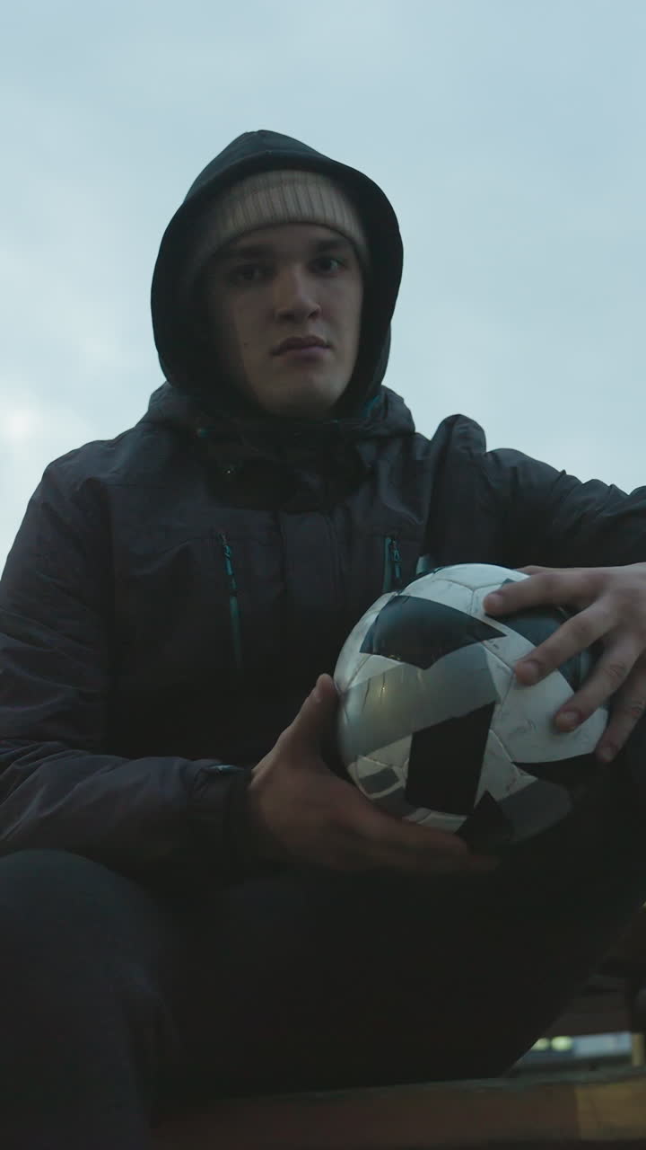 Young man in hoodie sitting on bench gripping soccer ball with intense focus, rotating it during urban training session, surrounded by modern residential buildings and evening lights
