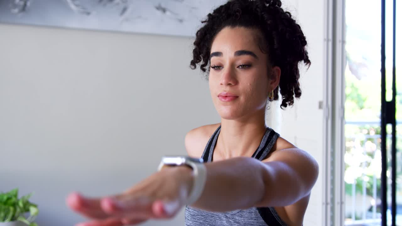 Donna afroamericana con canotta grigia che si allena mantenendo l'equilibrio con il braccio a casa, con uno smartwatch bianco.