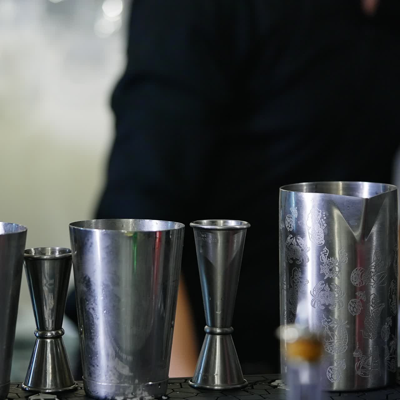 Metal cups and beavers used for making cocktails in the bar. Male in black shirt fills the metal jar with red beverage. Close up