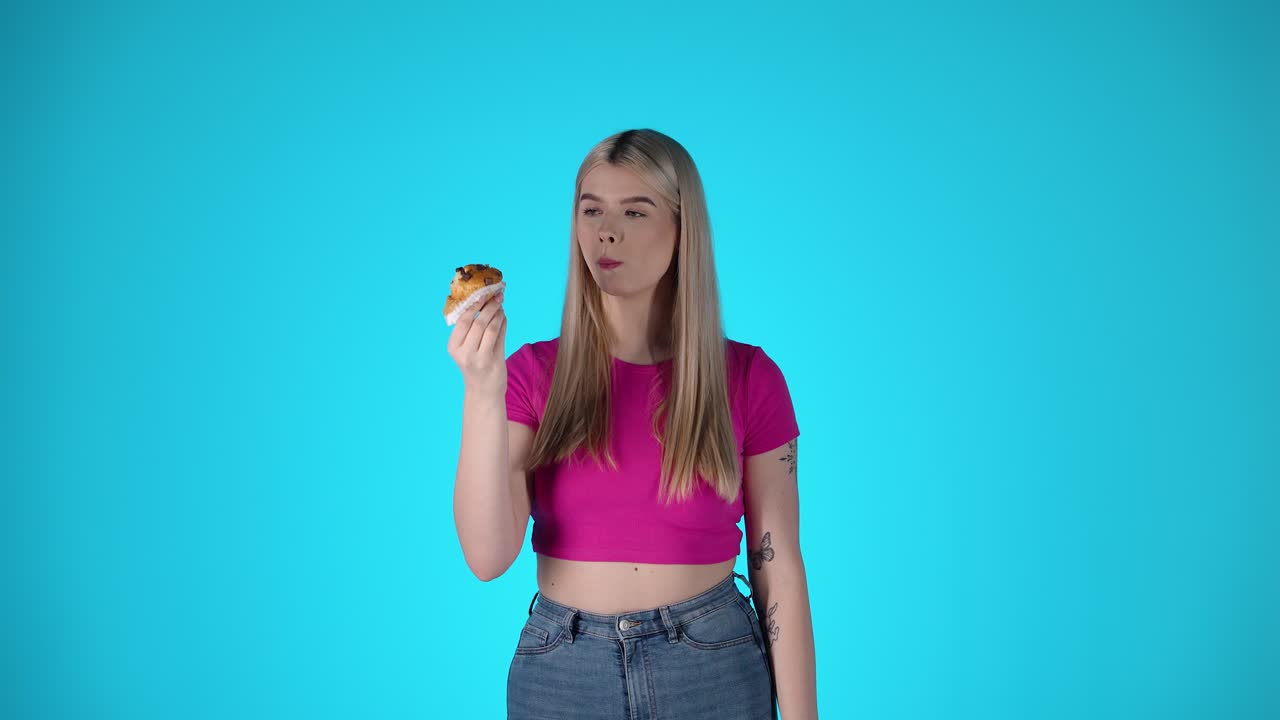 Pretty Young Blonde Woman Eating Pastry Against Blue Background, Studio Shot