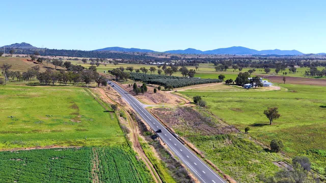 A drone glides over a rural two-lane road bordered by green fields, scattered trees, and distant hills under bright midday sunlight