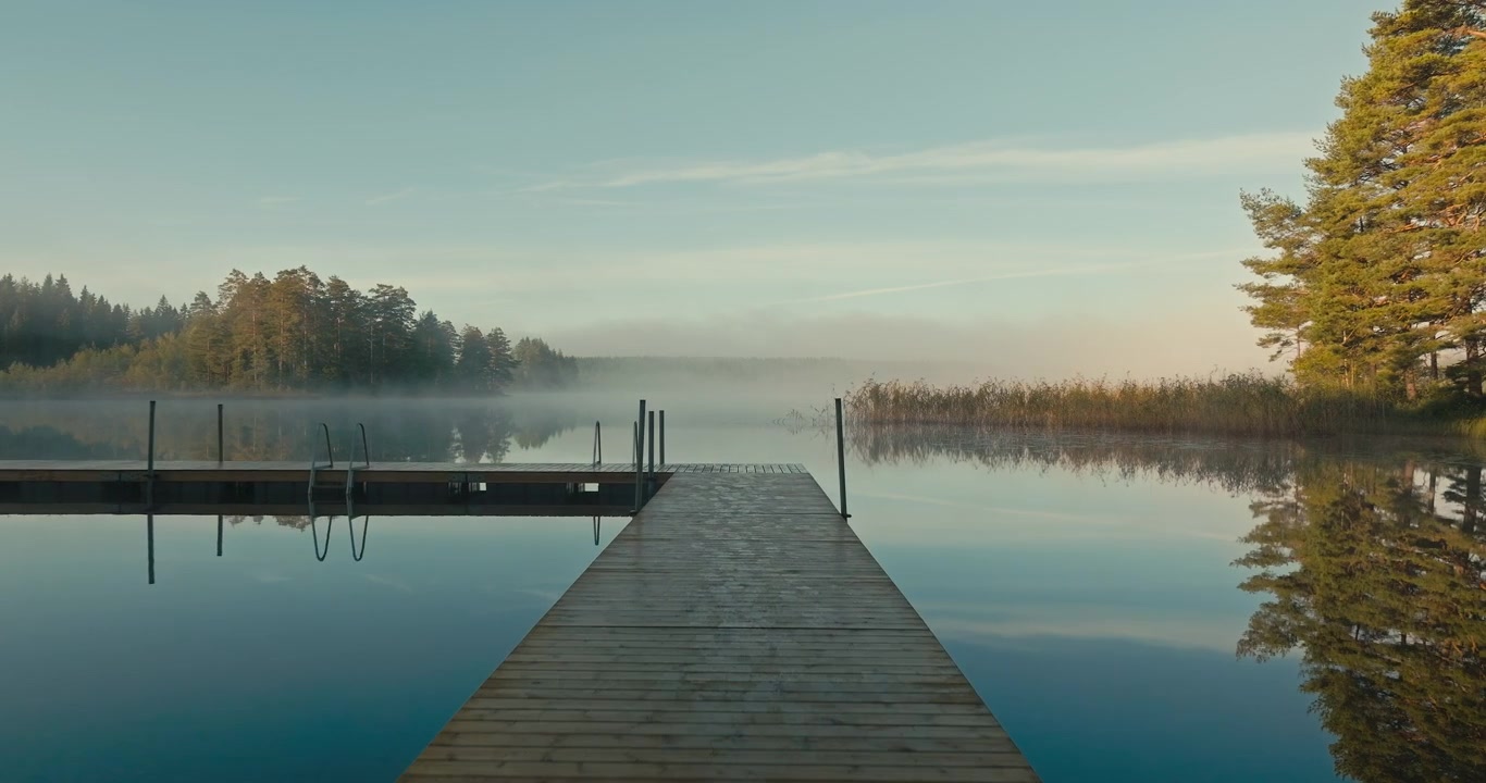 Drone flies over a wooden dock at a beautiful misty lake during sunrise, revealing calm water with stunning reflections of surrounding trees and peaceful natural scenery.
