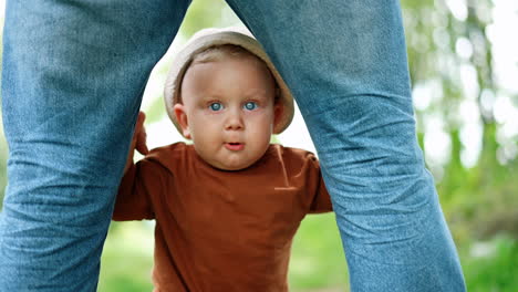 cute baby boy in a cap stands holding by his dad's legs. kid standing unsteadily learning to walk.