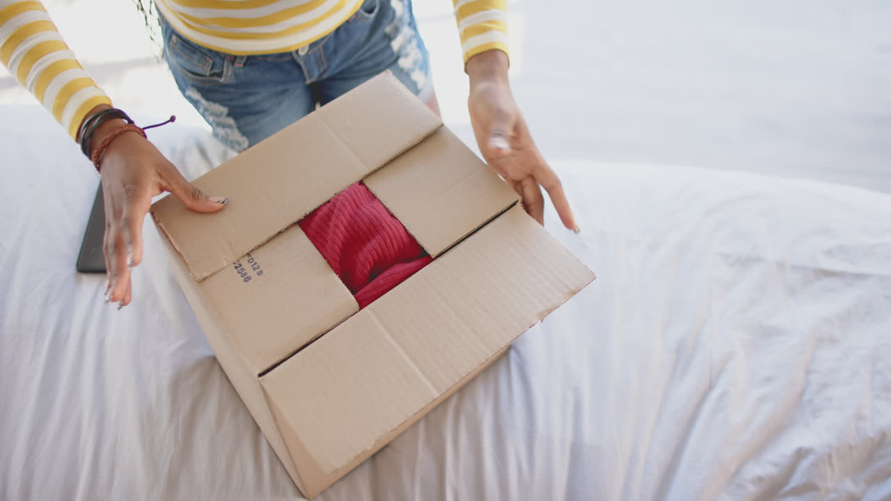 Opening cardboard box, African American woman unpacking red sweater on bed at home