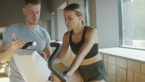 woman cycling in gym with help of personal trainer