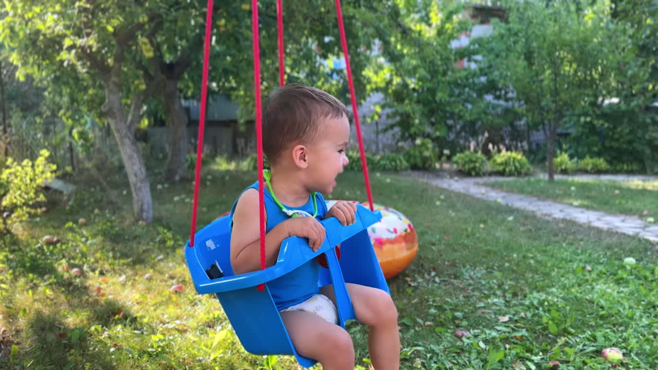 Excited Caucasian baby boy swinging cheerfully on the swing. Happy kid having fun time in the garden in summer.