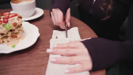 close view of woman's hand reaching toward cutlery wrapped in white napkin on wooden table. beside it sits a plate with stacked waffles topped with strawberries and cup of coffee in cozy cafe setting