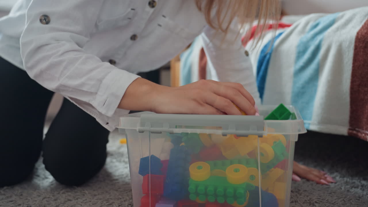 White mom tidying sons colorful blocks, kneeling on carpet beside plastic storage bin, hands reaching into box to sort large interlocking pieces, casual white shirt and black leggings, soft natural