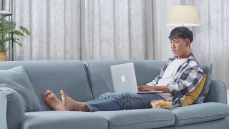 집에서 거실에서 소파에 누워있는 동안 노트북에 타이핑하는 간식과 함께 아시아 십대 소년의 몸이 가득 차 있습니다.
