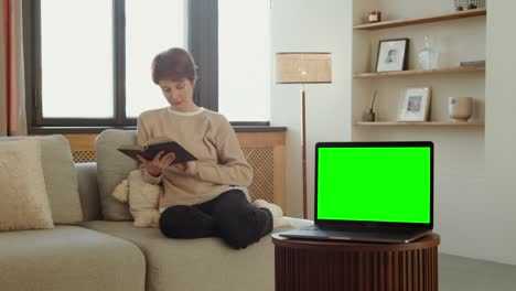 woman reading a book on a sofa with a green screen laptop