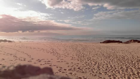 Golden-sand-beach-covered-with-footprints,-sunset-illuminates-clouds-over-ocean