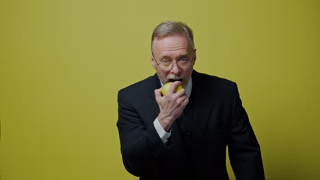 smiling senior businessman with fresh fruit. happy elderly man in elegant suit tries to eat apple and looking at camera. positive emotions.