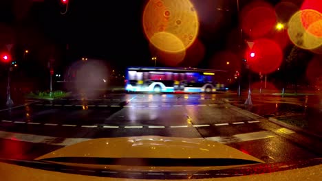 Stopping-at-the-night-at-a-red-traffic-light-by-car-while-a-bus-is-crossing-the-rainy-road