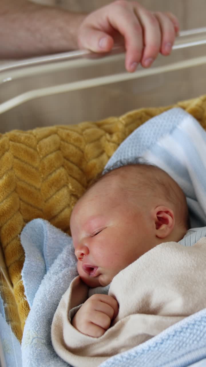 Un adorabile neonato caucasico dorme in una culla. Un uomo non identificato siede vicino alla culla del suo bambino appena nato. Vista dall'alto. Video verticale.