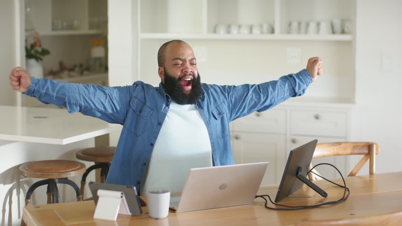 Uomo afroamericano che lavora in cucina con laptop e schermo, reagisce e si stira per la fatica.