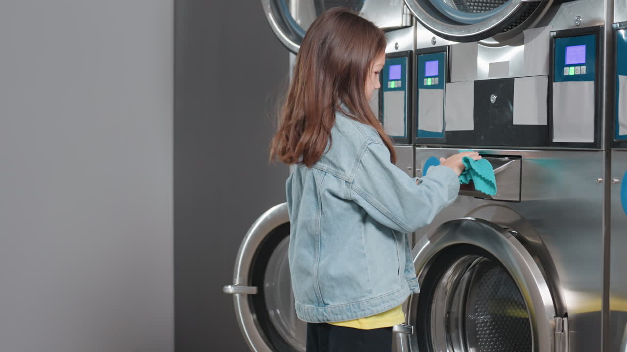 Back view girl in denim jacket scrubs washing machine surface using turquoise cloth inside laundromat, labels and screens visible, child cleaning routine maintenance, hygiene care, calm indoor scene