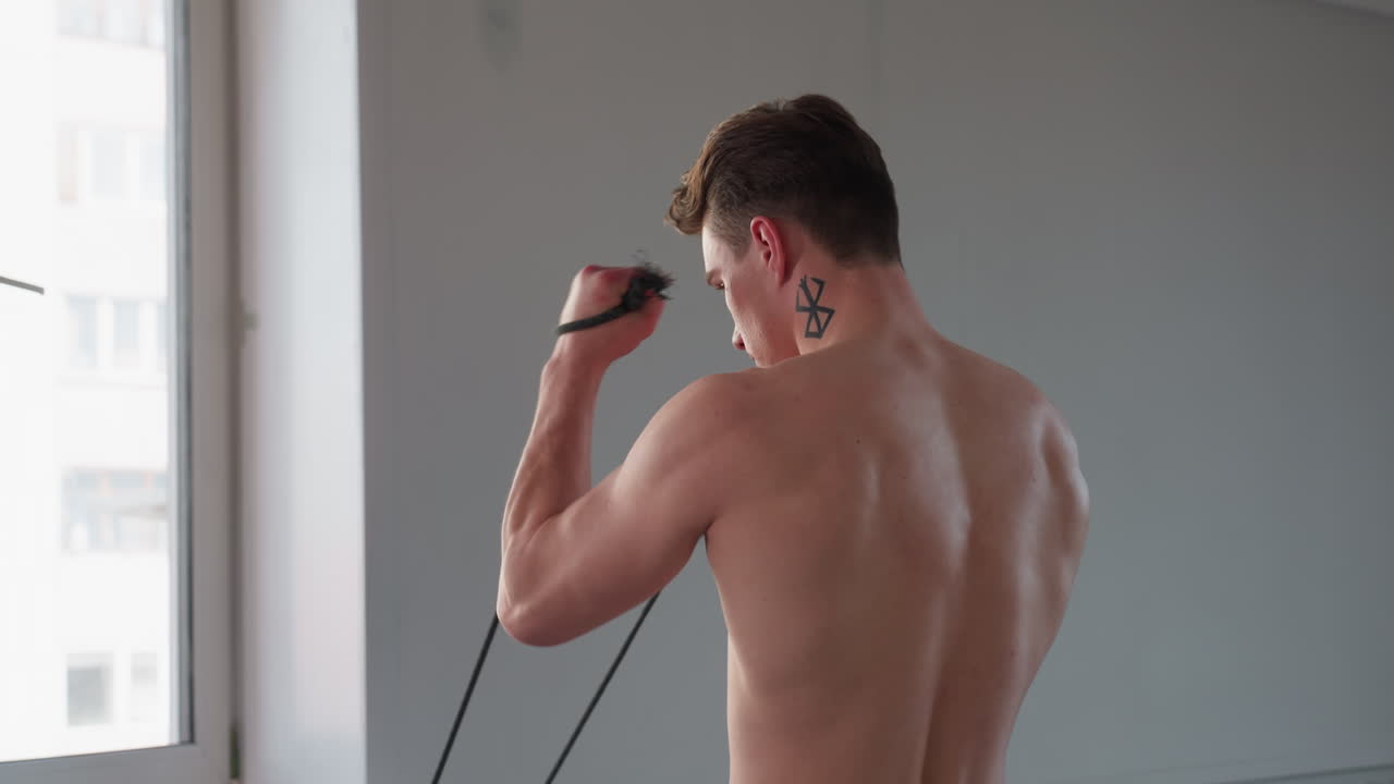 side view of shirtless young man with geometric neck tattoo pulling resistance rope near bright window in minimal room, sunlight casting soft shadows, office building exterior visible through glass
