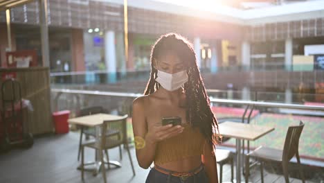 Young-black-woman-in-face-mask-uses-smart-phone-in-commercial-mall