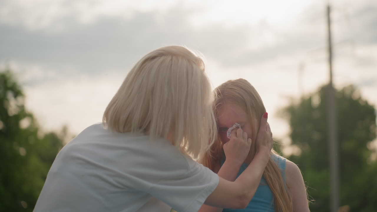 young girl wiping tears from cheek with tissue, sad expression visible, soft sunlight backlighting hair, blurred green background creating quiet emotional moment of vulnerability and resilience