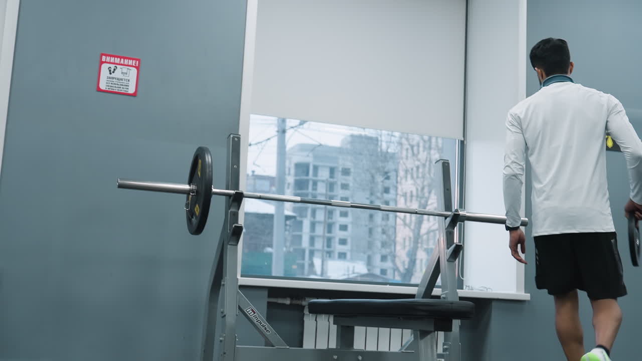 Young man carrying metal weight plate across gym floor to fix onto barbell on other side, with window showing residential building outside and rubber flooring, gym equipment