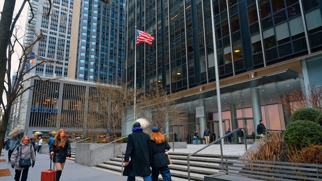 New York, USA, 15 aprile 2026: Bandiera americana fuori dagli edifici per uffici di Midtown Manhattan. Bandiera americana su un'asta vicino al 1345 di Avenue of the Americas, insegna Hilton, torri di vetro e pedoni.