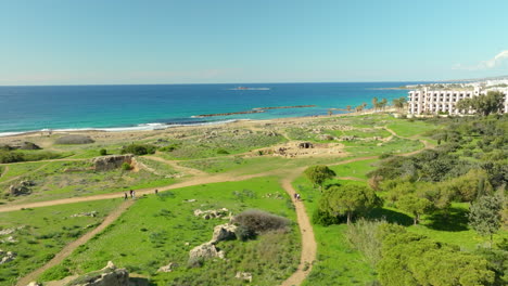 the tombs of the kings site is seen from a distance, with tourists walking among the ruins, set against the deep blue sea and green landscape, an emblem of cyprus's rich archaeological heritage