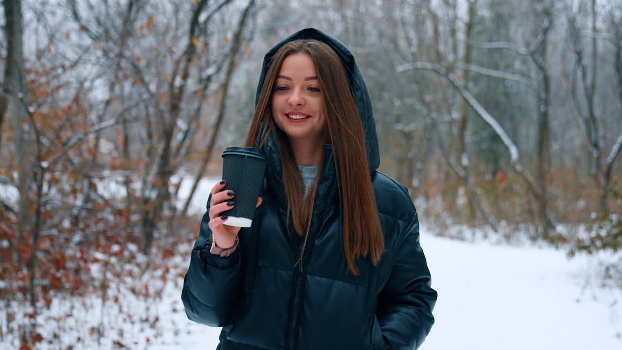 Happy relaxed woman with long brunette hair walks by the park in winter. Smiling lady with coffee cup in hands on the walk.