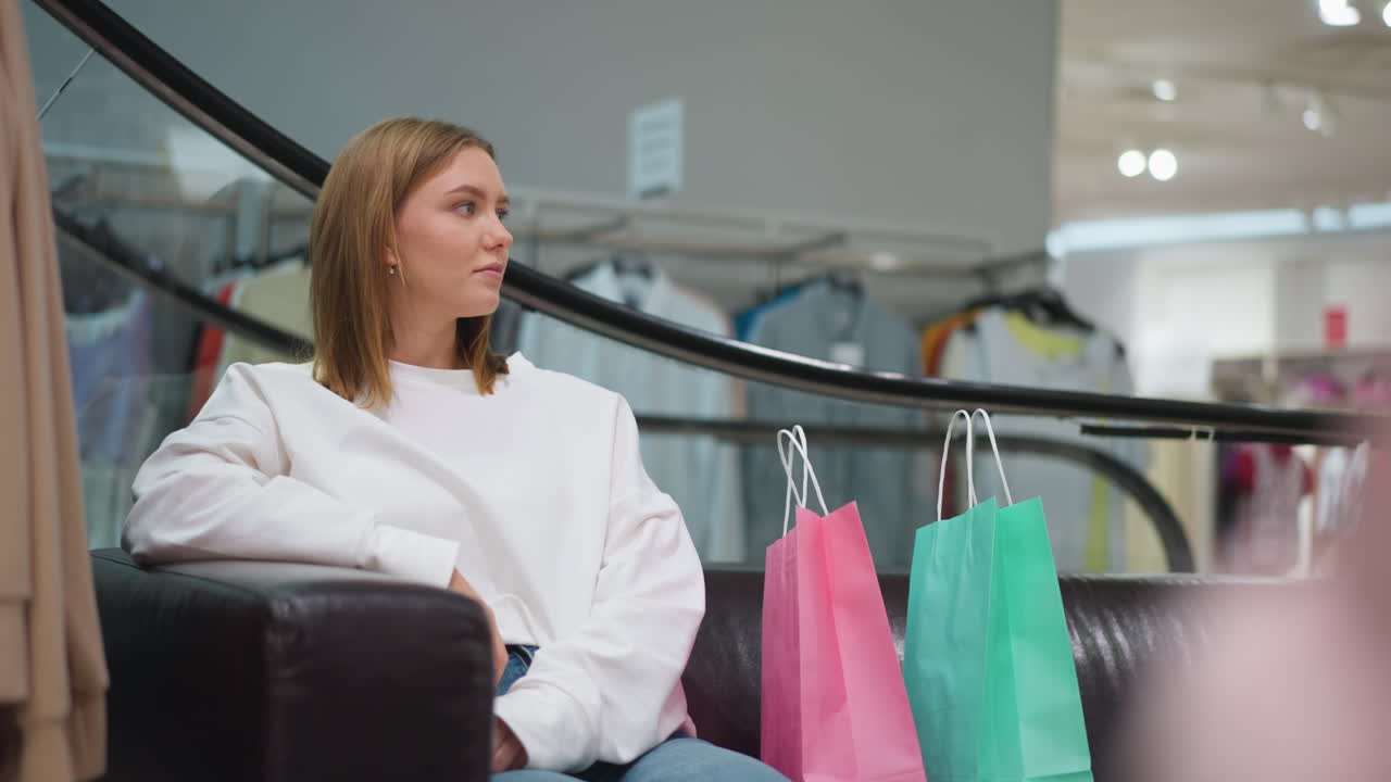 dama sentada en una silla de cuero en el centro comercial con bolsas de compras de colores colocadas a su lado, vestida casualmente con una camiseta blanca y vaqueros, rodeada de exhibiciones de ropa moderna bajo una iluminación suave
