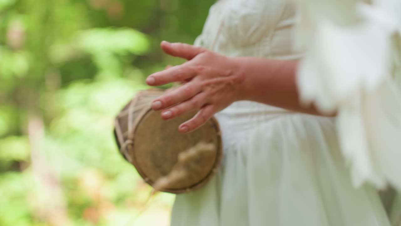 Close up of woman dressed in white gown with angel wings walking through forest while beating small hand drum, soft sunlight touching her hands, creating magical peaceful moment