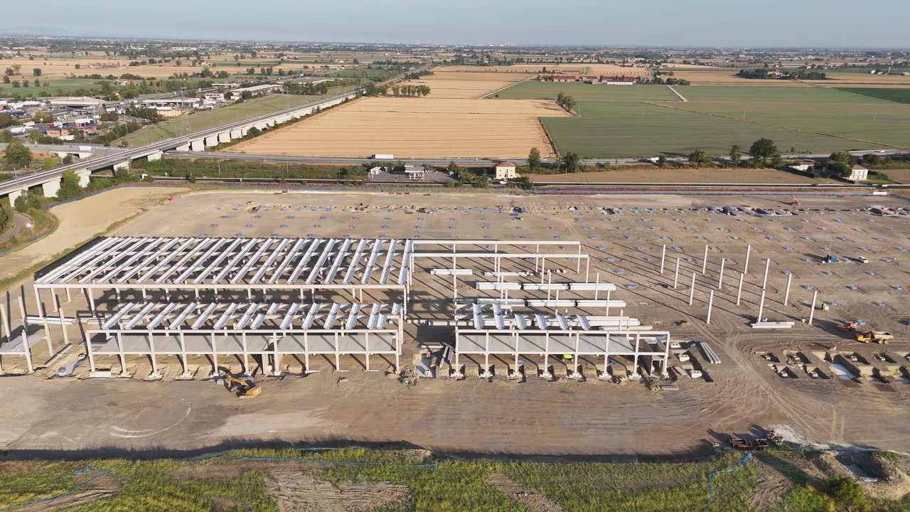 Costruzione di un parco logistico a Barabasca vicino all'uscita dell'autostrada A1 di Fiorenzuola d'Arda, con strutture in acciaio per magazzini, cantieri edili e terreni agricoli coltivati della Pianura Padana, ripresa con drone.