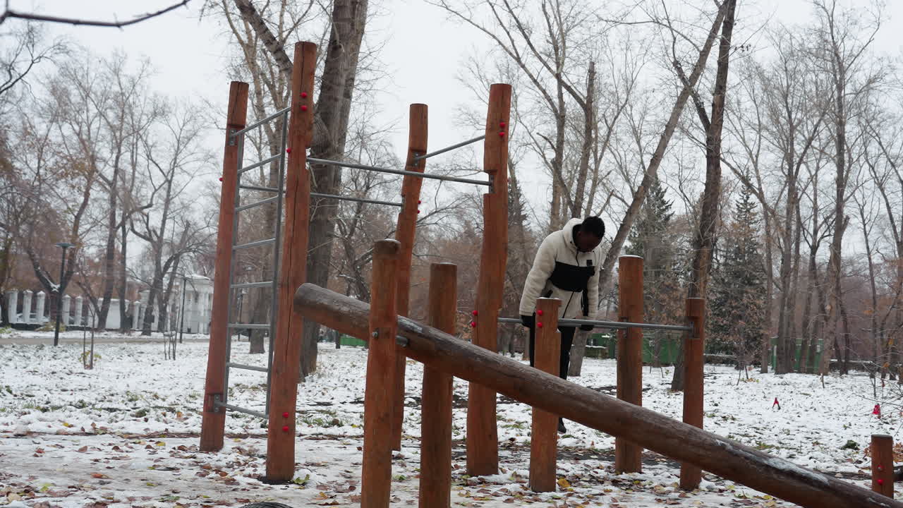 흑인 남자가 겨울 동안 야외 운동 장비에 올라서 눈으로 인 땅과 배경에 벌거벗은 나무로 둘러싸인 밀어붙이는 운동을 수행합니다.