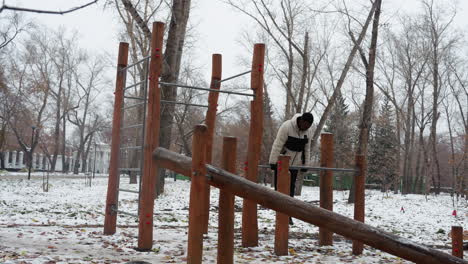 hombre negro sube en equipos de entrenamiento al aire libre durante el invierno, realizando ejercicios de empuje mientras cae, rodeado de tierra cubierta de nieve y árboles desnudos en el fondo