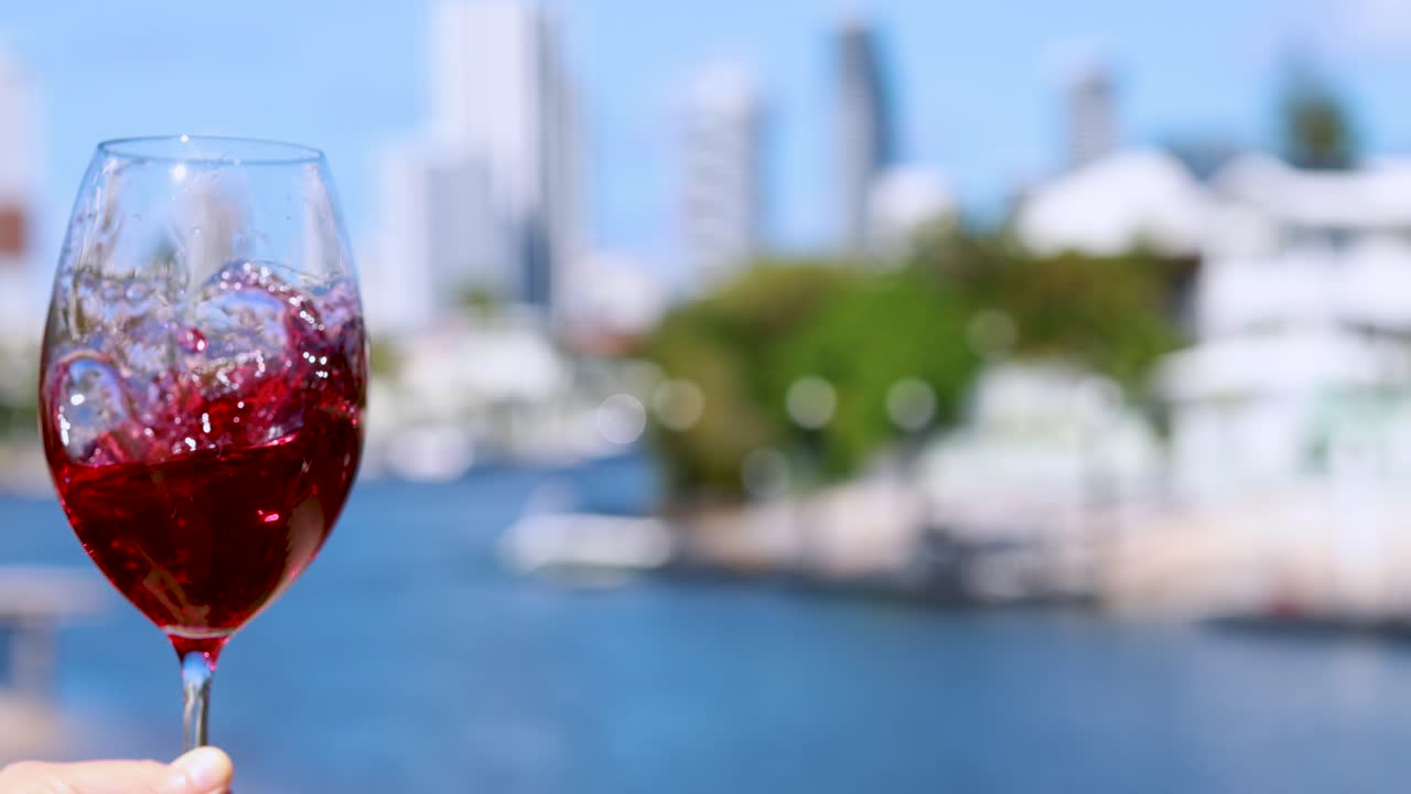 A hand gently swirls a glass of red wine against a bright, sunlit waterfront cityscape. Shallow depth of field highlights the swirling motion and vibrant colors