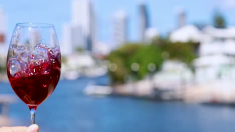 a hand gently swirls a glass of red wine against a bright, sunlit waterfront cityscape. shallow depth of field highlights the swirling motion and vibrant colors