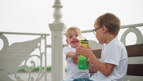 a younger sibling with chocolate-stained mouth hands a water bottle to his older brother, as he sticks his tongue out