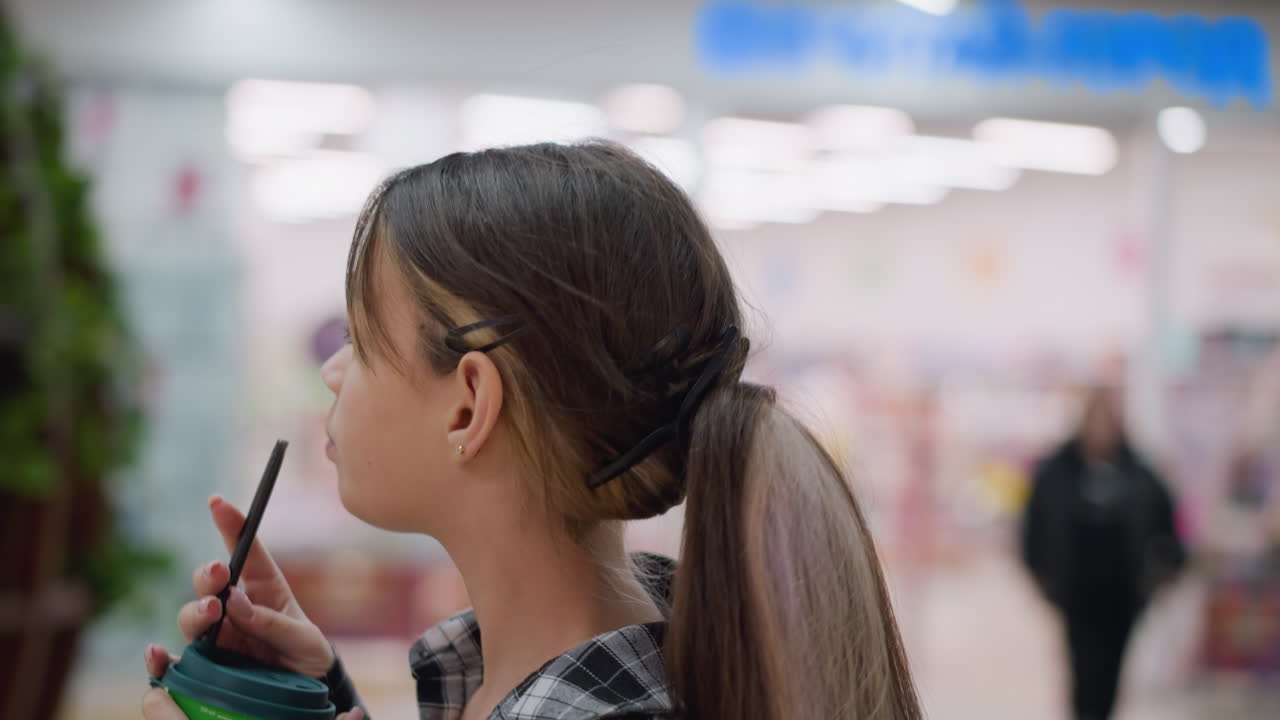 현대적인 실내 공간에서 그녀의 좌석을 향해 부드럽게 걸어가면서 주스를 마시는 어린 소녀의 클로즈업 뷰, 흐릿한 배경 조명과 멀리서 부드럽게 초점을 맞추지 않는 인물이 특징입니다.