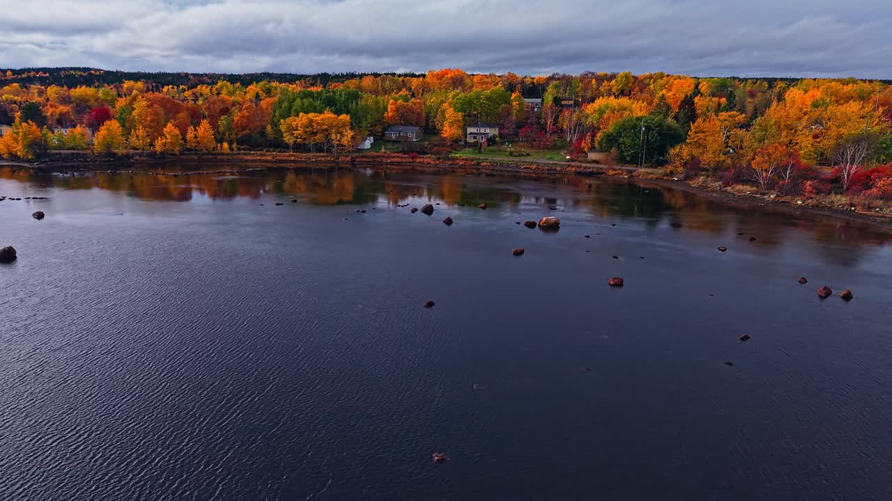 Sweeping aerial reveals a narrow road dividing a tranquil bay with rocky shallows, while brilliant fall foliage and houses line Port Blandford’s shore