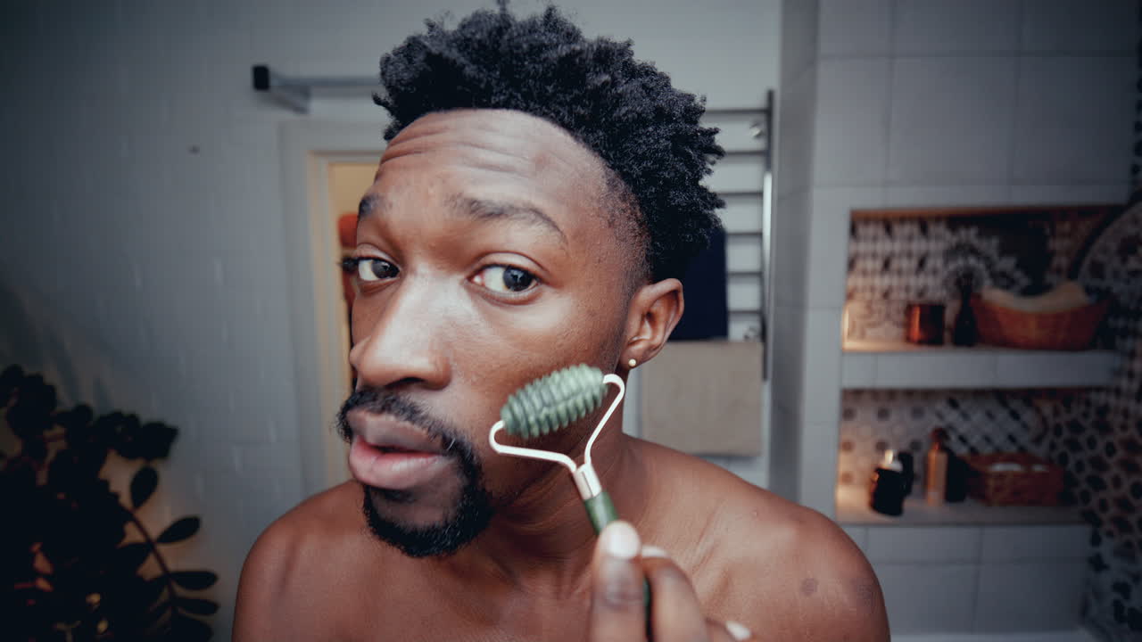 Giovane uomo di colore che si massaggia il viso con un rullo di giada in bagno.
