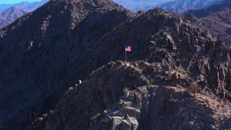 Toma-Aérea-De-La-Bandera-Americana-En-La-Cima-De-La-Montaña-Con-Fondo-De-Montaña