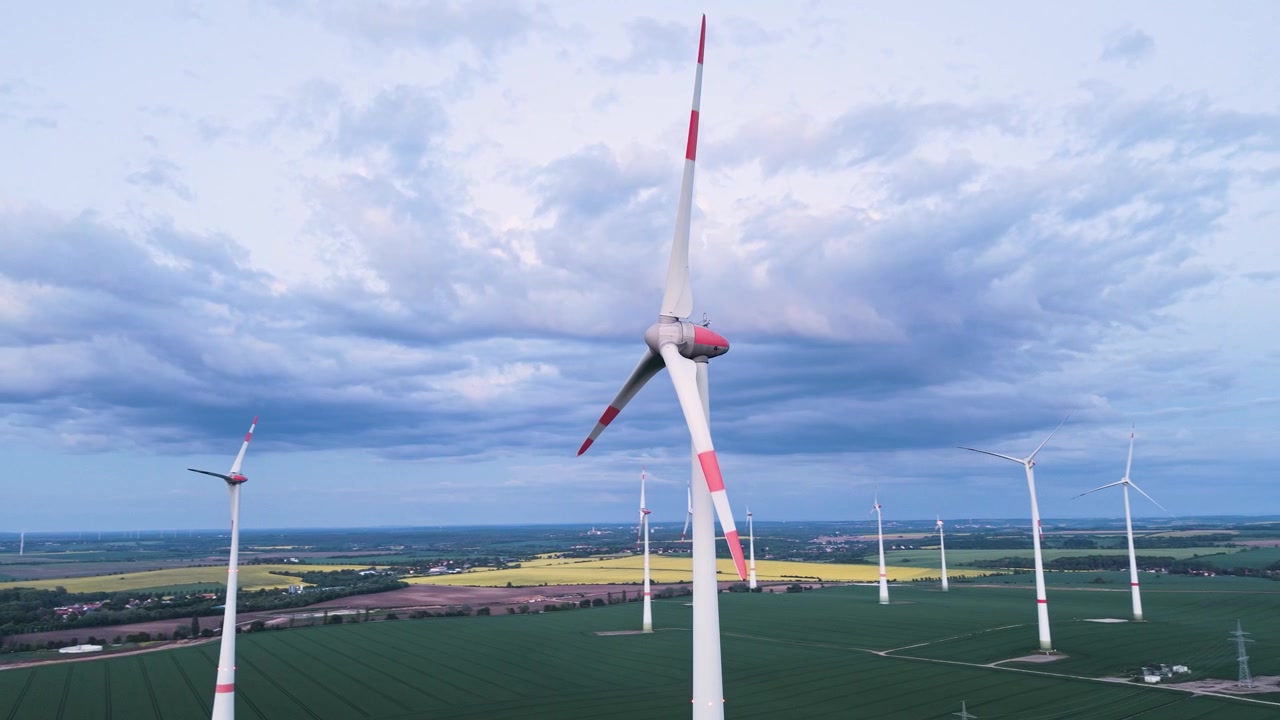 Droneflyvning over en stor vindmøllepark på landet i Tyskland med tårnhøje turbiner over grønne landbrugsmarker under dramatiske, skyfyldte aftenshimle, der fremviser produktion af vedvarende energi i UHD
