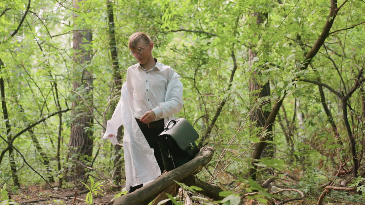 Botany student in white shirt pulling off white coat in lush green forest near black leather bag on tree branch, surrounded by foliage, branches, trunks, and natural sunlight