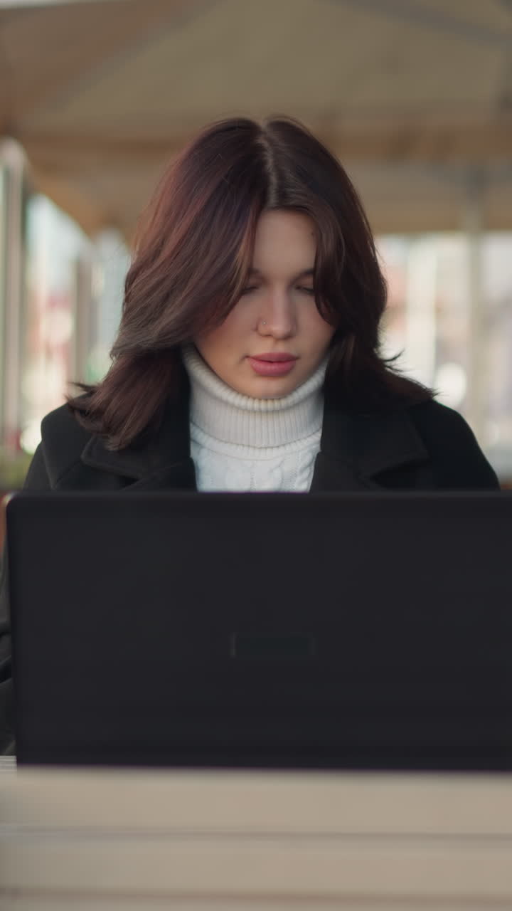 dama trabajando en una computadora portátil en un entorno urbano, con un elegante abrigo negro, con un pilar negro decorativo cerca, un edificio de ladrillo rojo borroso en el fondo, y una figura borrosa con vegetación en la distancia