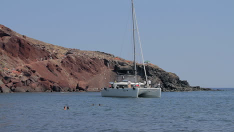 a catamaran sails by the red beach of santorini