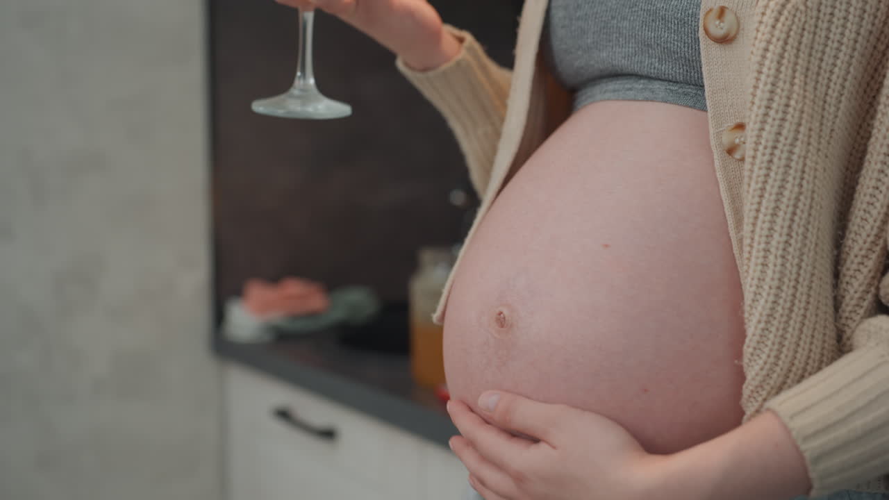 Donna incinta che tiene un bicchiere di succo, scena in cucina che mostra la futura mamma in un cardigan accogliente che tocca la pancia nuda con dolcezza, primo piano della texture della pelle e del pancione arrotondato, luce calda del mattino e calma