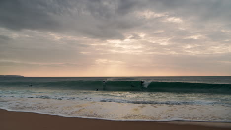 Toma-Amplia-Desde-La-Playa-Hasta-El-Océano-Durante-La-Puesta-De-Sol,-Donde-Los-Surfistas-Están-Surfeando-Y-Esperando-Una-Ola