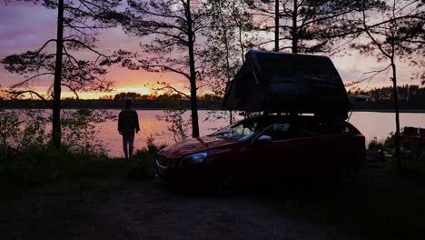 woman stands by swamp lake during colorful sunset, rooftop tent camping, latvia
