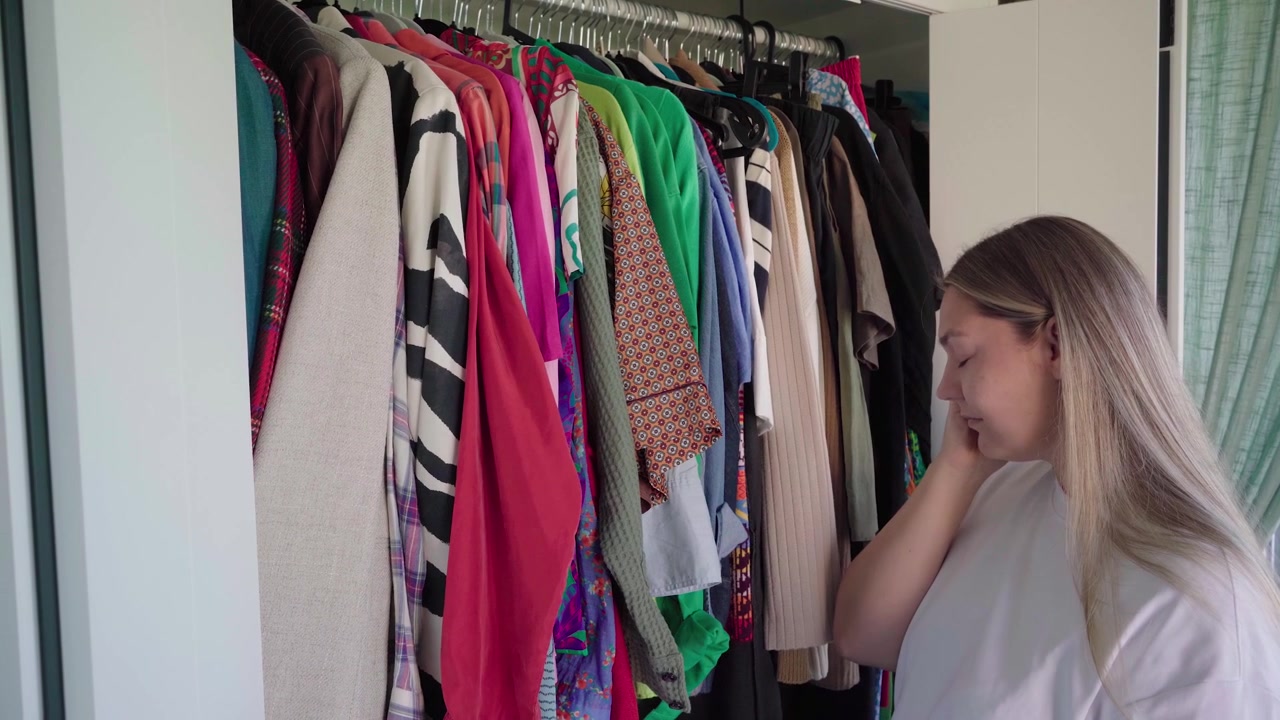 Mulher estressada está diante de um guarda-roupa cheio, tocando as têmporas enquanto uma dor de cabeça se intensifica ao tentar escolher o que vestir
