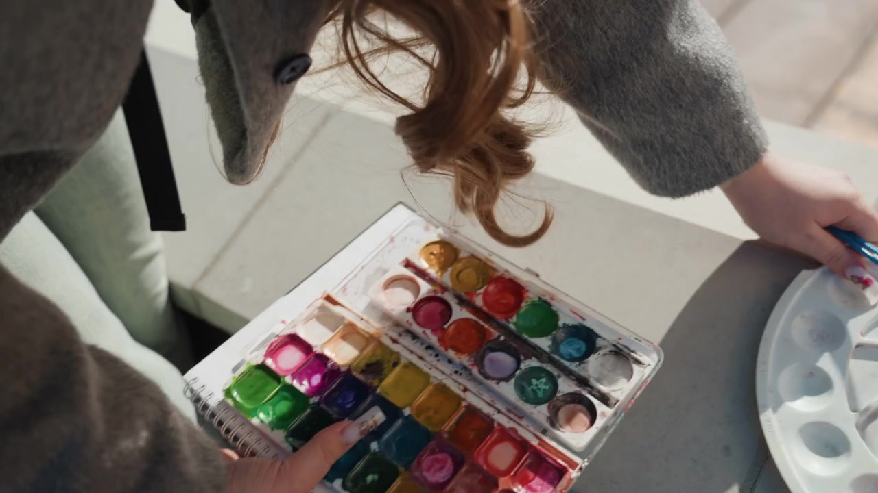 Nahaufnahme einer Aquarellpalette und eines Pinsels auf dem Dach. Eine junge Frau ordnet Farben auf einem Betonsims an, Sonnenlicht hebt lockiges Haar und den Ärmel eines Wollmantels hervor, mintfarbene Hose sichtbar, Hände wählen leuchtende Farbnäpfchen aus.