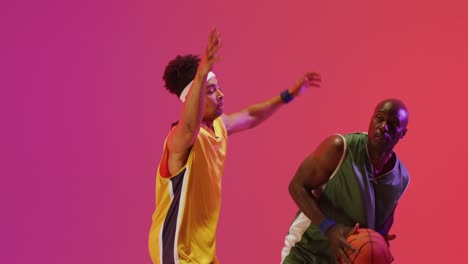 Video-of-two-diverse-male-basketball-players-playing-with-ball-on-pink-background